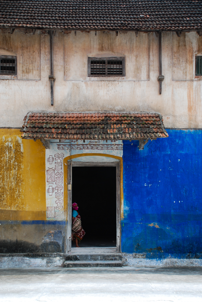 A picture of a yellow and blue facade with a tall doorway. There is a lady peering out of it. Travel, minimalist photography by Yuvan Kumar.