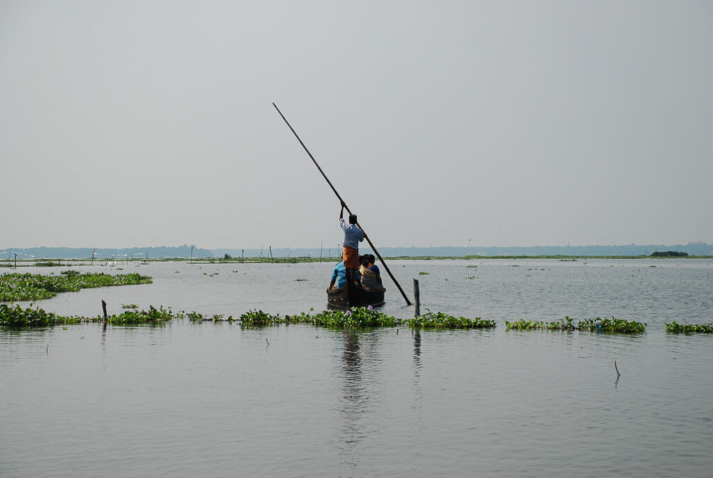 A man rows a boat full of people using a long bamboo stick as an oar on the Vembanad River