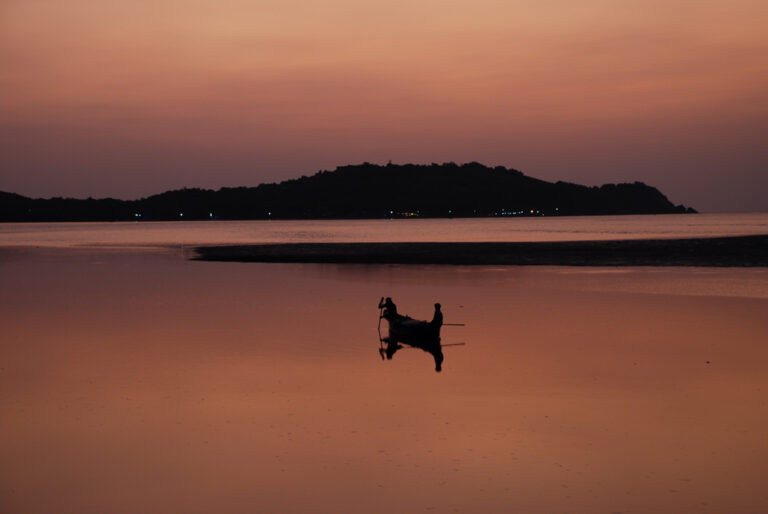 Two fishermen are pictured in their boat in a bay at sunset in Siolim, Goa, in Southern India. Th photograph was originally clicked by Yuvan Kumar and can be categorised as travel photography, minimalism, silhouettes. The colours are pink, orange.