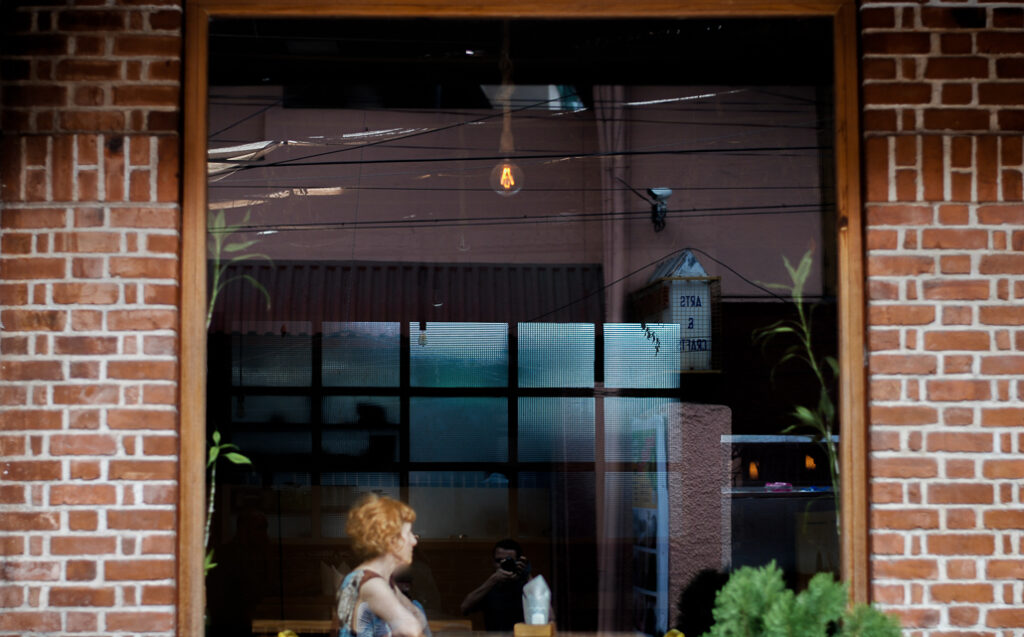 A Caucasian woman with ginger hair is sitting in a cafe/restaurant in Kochi, Kerala, India. There is a light bulb hanging over her and the facade of the building is built in red brick. She is sitting alone, perhaps a tourist, a traveller on vacation to South India. This photograph is an original taken by journalist and photographer Yuvan Kumar.