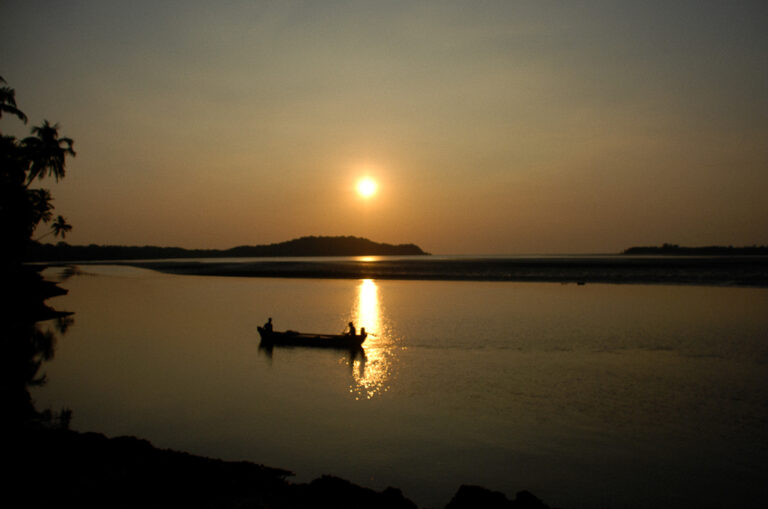 Two fishermen are pictured in their boat in a bay at golden hour, sunset in Siolim, Goa, in Southern India. Th photograph was originally clicked by Yuvan Kumar and can be categorised as travel photography, minimalism, silhouettes.