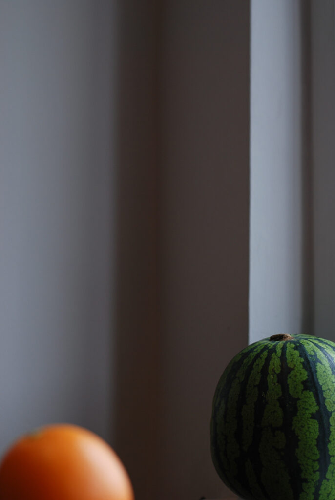 A still photograph of an orange and a green watermelon leaning against a window. This photograph is minimalism, abstract art, still life, indoor photography, COVID art. Originally clicked by Yuvan Kumar.